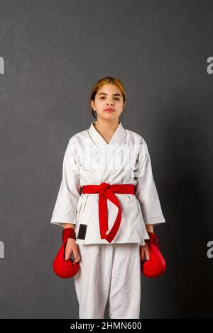 A young karateka girl in a white kimono and red competition outfit trains and performs a set of exercises against a gray wall Stock Photo