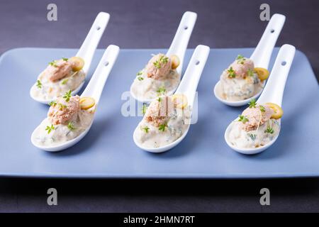 Cold appetizer made from cod liver, cod caviar, olives, cucumber and microgreens on serving spoons. Traditional cold dish. Close-up, blue plate, black Stock Photo