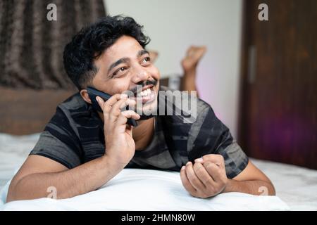Happy young man talking on mobile phone by lying on bed at home - concept of relationships, chatting with girlfriend and communication. Stock Photo