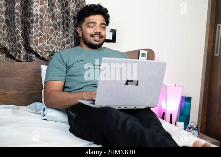 Young man busy working online using laptop at bedroom - concept of work from home, relaxation and new normal during covid-19 pandemic. Stock Photo