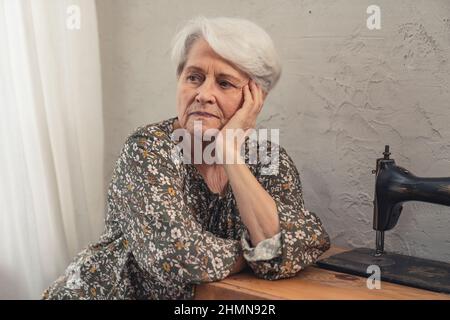 upset caucasian pensioner in her 70s sitting alone next to the sewing machine and propping up her wrinkled chick. High quality photo Stock Photo
