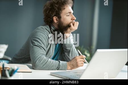 Remote working pensive young man working at home. Serious young freelancer man sitting at the desk and using laptop at home office and taking notes. Work from home concept.  Stock Photo