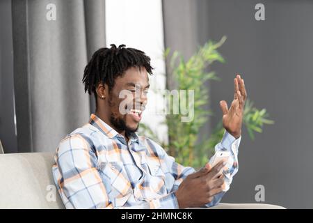Happy laughing African American student use smartphone having video call or watching online videos. Portrait of happy man gesturing looking at smartphone sitting on the sofa. Social media concept.  Stock Photo