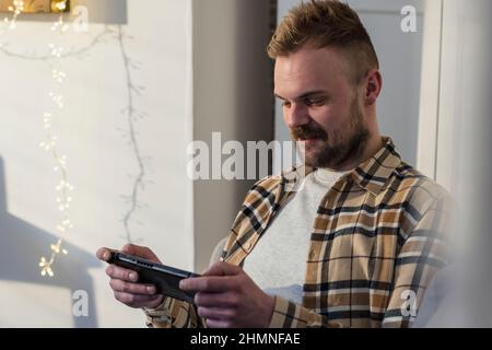 A side-view shot of a young man sitting on his sofa in his living room relaxing, he is using his smart phone. Stock Photo