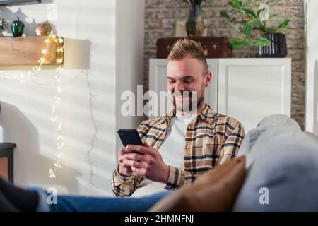 A front-view shot of a young man lying on his sofa in his living room relaxing, he is using his smart phone. Stock Photo