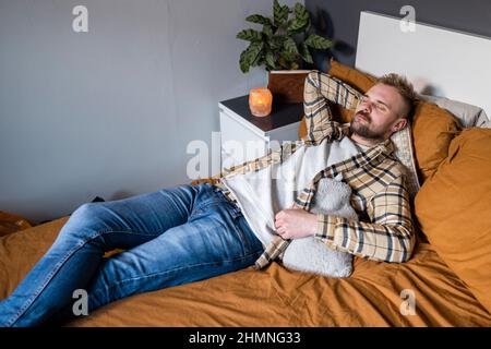 A high angle view shot of a young man lying on his bed in his bedroom relaxing, he has his eyes closed and a hot water bottle by his side. Stock Photo