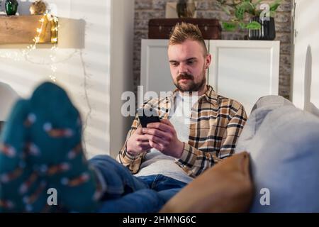 A front-view shot of a young man lying on his sofa in his living room relaxing, he is using his smart phone. Stock Photo