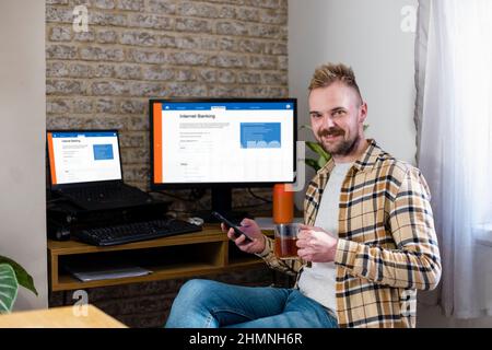 A man is sitting holding his phone by his desk while looking at the camera with a smile. Internet banking is displayed on his work screens. Stock Photo