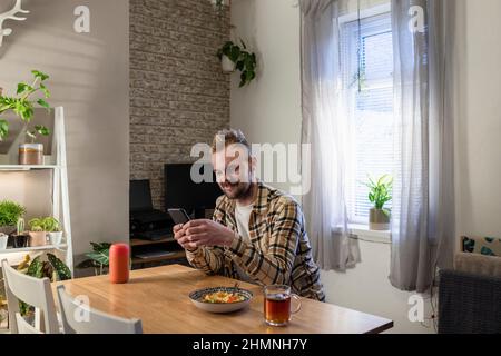 A side-view shot of a young man sitting at a table in his living room relaxing, he is using his smart phone and he has a plate of pasta near by. Stock Photo