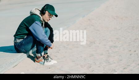 a handsome middle-aged man, wearing modern casual clothes, listens to music using a pair of headphones sitting on the promenade, next to the sand of a Stock Photo