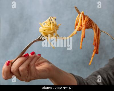 A woman's hands hold two forks with spaghetti in white on one side and spaghetti with tomato sauce on the other Stock Photo