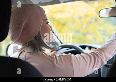 Charming blond asian woman driving car. Mature asian woman in glasses looking at the mirrow holding a steering wheel. Blond middle aged asian woman in the car. Woman on the road. Business concept.  Stock Photo