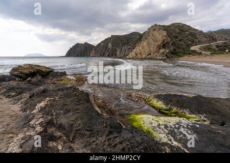 A secluded sandy beach on a wild mountainous coastline with colorful rocks and algae in the foreground Stock Photo