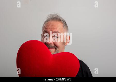 Handsome middle-aged gray-haired and bearded man holding a red heart, isolated gray background. Valentine's day concept Stock Photo