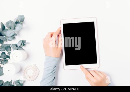 Female hands using tablet pc with blank display next to pearl necklace, candles and eucalyptus branches on white table, top view. Stock Photo