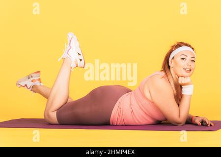 Beautiful young woman with an oversized body resting after workout lying on fitness mat smiling looking at camera, yellow background. Stock Photo