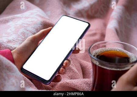 Woman sitting on bed and using mobile phone with blank white desktop screen. Mockup image of hand holding smartphone. Stock Photo