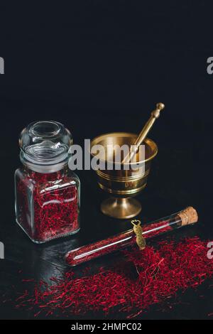 Raw organic red dried saffron spice on wooden background in vintage metal brass mortar with pestle, glass jar and tube Stock Photo