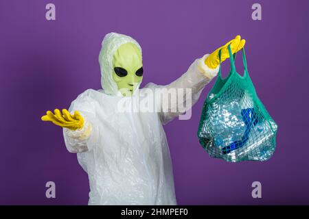 Funny man in an alien mask in a white suit holds a mesh bag with plastic bottles and spreads his hands on a purple background, waste sorting concept Stock Photo