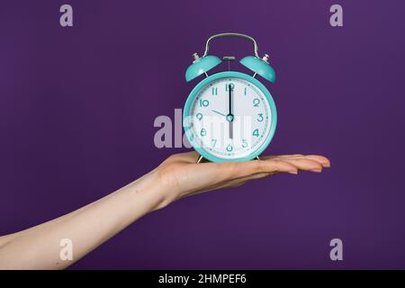Woman's hand holds a turquoise alarm clock in the palm of her hand on a purple background Stock Photo