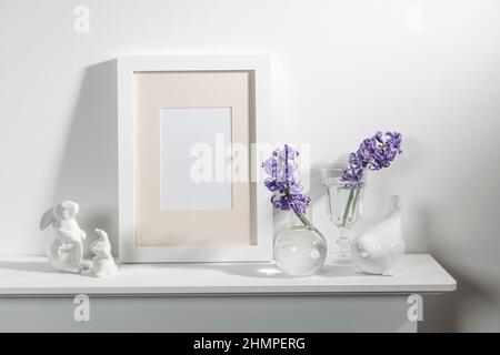 White hyacinth in a large porcelain bowl, books, figurines of hares and a bird, an empty photo frame are on the fireplace against the white wall. Layo Stock Photo