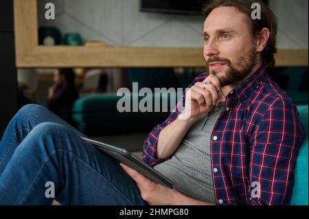 Pensive handsome bearded Caucasian man in casual denim and checkered shirt holding a digital tablet and looking thoughtfully away, sitting at home env Stock Photo