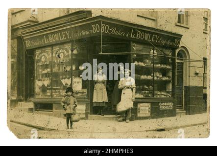 Original, charming post WW1 era shop front of L. Bowley Baker and Confectioner. A classic independent corner shop. Outside posing for a photograph are the baker & female shop assistants. The baker wears an apron, hat and has a built up shoe, invalided from the war?. A cute small girl wearing a cloche hat stands in the street. circa 1920, 1921, U.K. Europe. Stock Photo