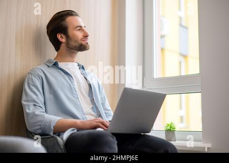 Cheerful caucasian guy sits on sofa in modern living room alone and using trendy laptop for net searching, web surfing or online learning Stock Photo