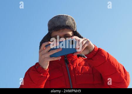 A young man of European appearance in a red jacket and a winter hat takes pictures from the phone in Stock Photo