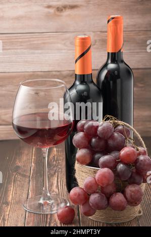 The photo shows two black wine bottles with orange caps. A glass of red wine half filled. Basket with large pink grapes. Composition woody background. Stock Photo