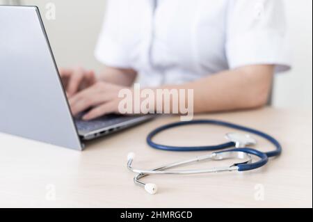 Unrecognizable female doctor in uniform uses a laptop. Faceless medical worker writes a report on the computer at the desk. Cropped. Stock Photo