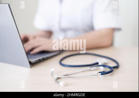 Unrecognizable female doctor in uniform uses a laptop. Faceless medical worker writes a report on the computer at the desk. Cropped. Stock Photo