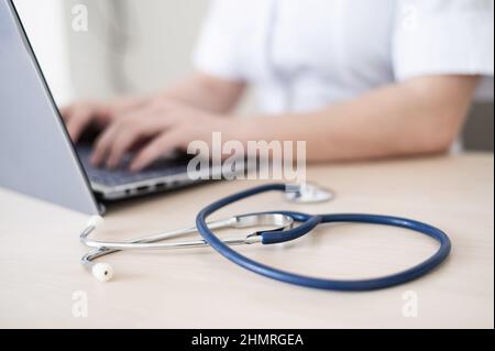 Unrecognizable female doctor in uniform uses a laptop. Faceless medical worker writes a report on the computer at the desk. Cropped. Stock Photo