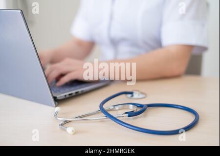 Unrecognizable female doctor in uniform uses a laptop. Faceless medical worker writes a report on the computer at the desk. Cropped. Stock Photo