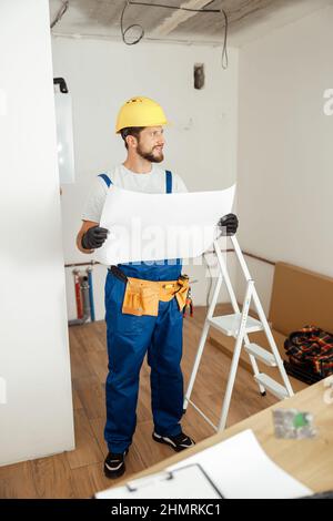 Full length shot of thoughtful contractor in overalls and hard hat looking away while studying blueprint, standing in new apartment Stock Photo
