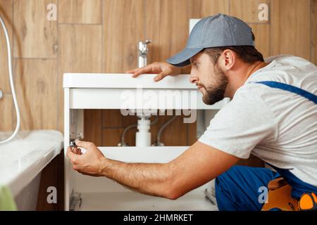 Concentrated plumber, male worker in uniform fixing sink and water pipe in the bathroom Stock Photo