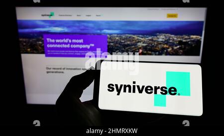 Person holding mobile phone with logo of US telecom company Syniverse Technologies LLC on screen in front of web page. Focus on phone display. Stock Photo