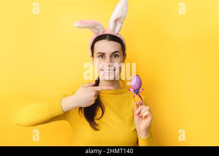 Portrait of smiling beautiful young brunette female wears fluffy bunny ears, pointing on small colored eggs, wears sweater, posing isolated on yellow Stock Photo