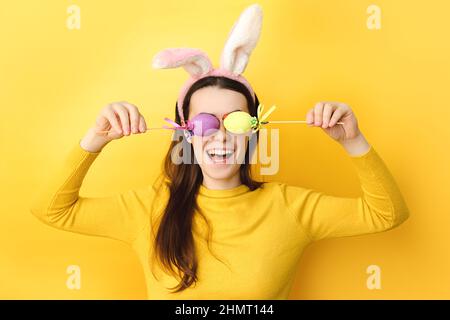 Funny young caucasian woman covers eyes with two Easter eggs, wears fluffy bunny ears, dressed in sweater, model on yellow background with free space Stock Photo