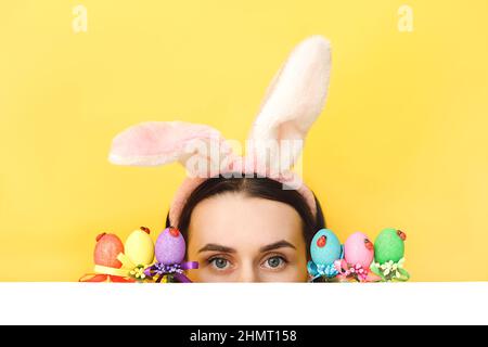 Close up portrait of fun young woman wears bunny ears, hides face near Easter eggs, looks mysteriously from into the camera , isolated on yellow color Stock Photo