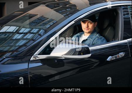 Portrait of male driver in cap sitting in electric car. Handsome young man driving modern electric automobile. Concept of transport and electro mobility. Stock Photo