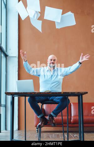 Dissatisfied mature businessman at office desk throwing paper documents in air  Stock Photo