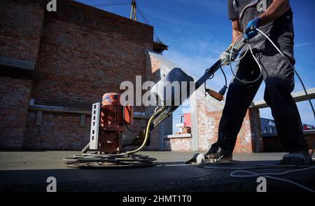 Cropped picture of man laborer in work overalls using power trowel machine while screeding floor in new building. Male worker finishing concrete surface with troweling machine at construction site. Stock Photo