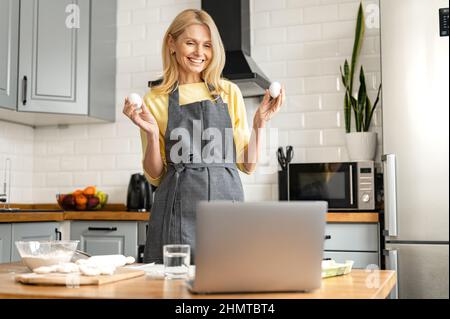Positive middle-aged woman in apron holding two eggs going to prepare a dough and streaming it in social media, female cooking and streaming process by laptop, conducts cooking video class Stock Photo