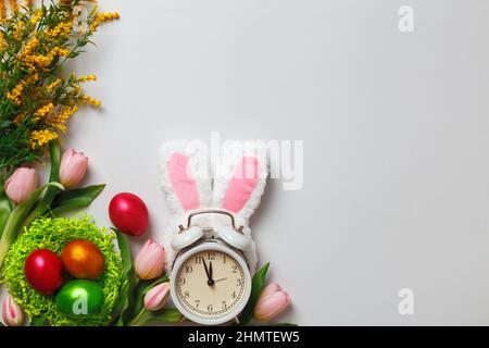 Easter composition. Multicolored pearl eggs in a nest alarm clock rabbit ears flowers tulips on a gray background with copy space. Stock Photo