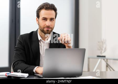 Glad male entrepreneur looking at the camera. Cheerful caucasian man wearing formal wear sitting at the table and using laptop at modern office Stock Photo