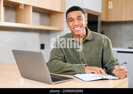 Cheerful man using a laptop for work while sitting and answering emails. Guy making notes at the notebook and looking at the camera with pleasure smile Stock Photo