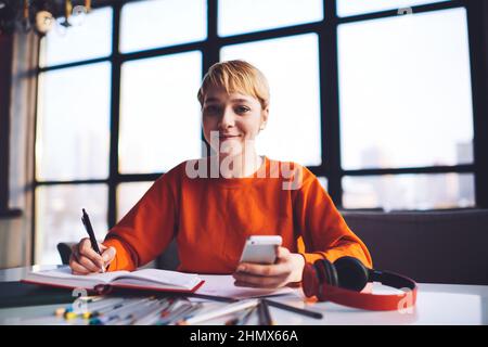 Happy female student writing in notepad and using smartphone in workspace Stock Photo