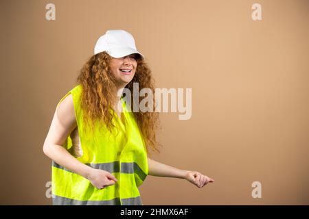 A red haired woman in a yellow vest and and a cap on her head posing with a smile over orange background and copy space. Stock Photo