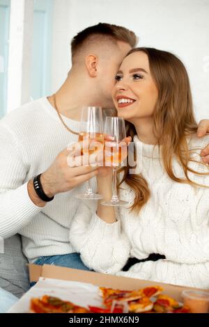 Two tall glass glasses with wine in hands of joyful couple in love. Young people in white knitted sweaters hug, man whispers something tender in ear Stock Photo